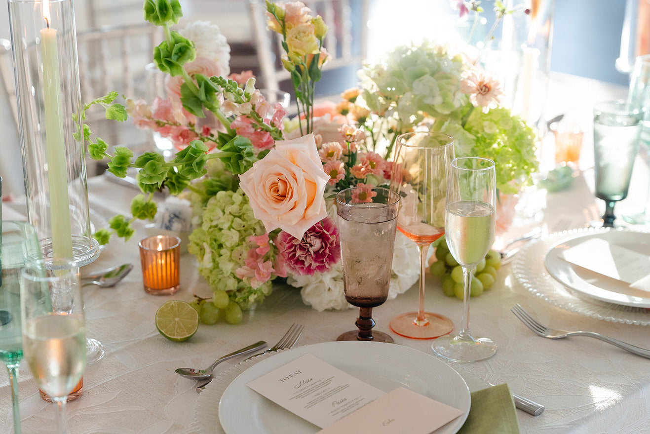 A beautifully styled wedding table setting featuring a floral centerpiece with pink, peach, and green flowers, white plates, green napkins, and tinted glassware on a white tablecloth.